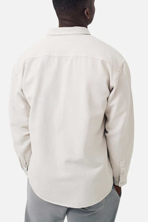 The image displays a person from the back wearing Industrie The Portland Shirt, a light beige cotton-linen overshirt with a relaxed fit, buttoned cuffs, and classic collar. They pair it with light gray pants against a plain white background.