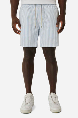 A person stands facing forward, wearing light blue and white striped Industrie The Princeton Bahama Shorts featuring a drawstring waist, paired with white sneakers. These shorts provide a comfortable fabric blend ideal for any casual day. The background is plain white.
