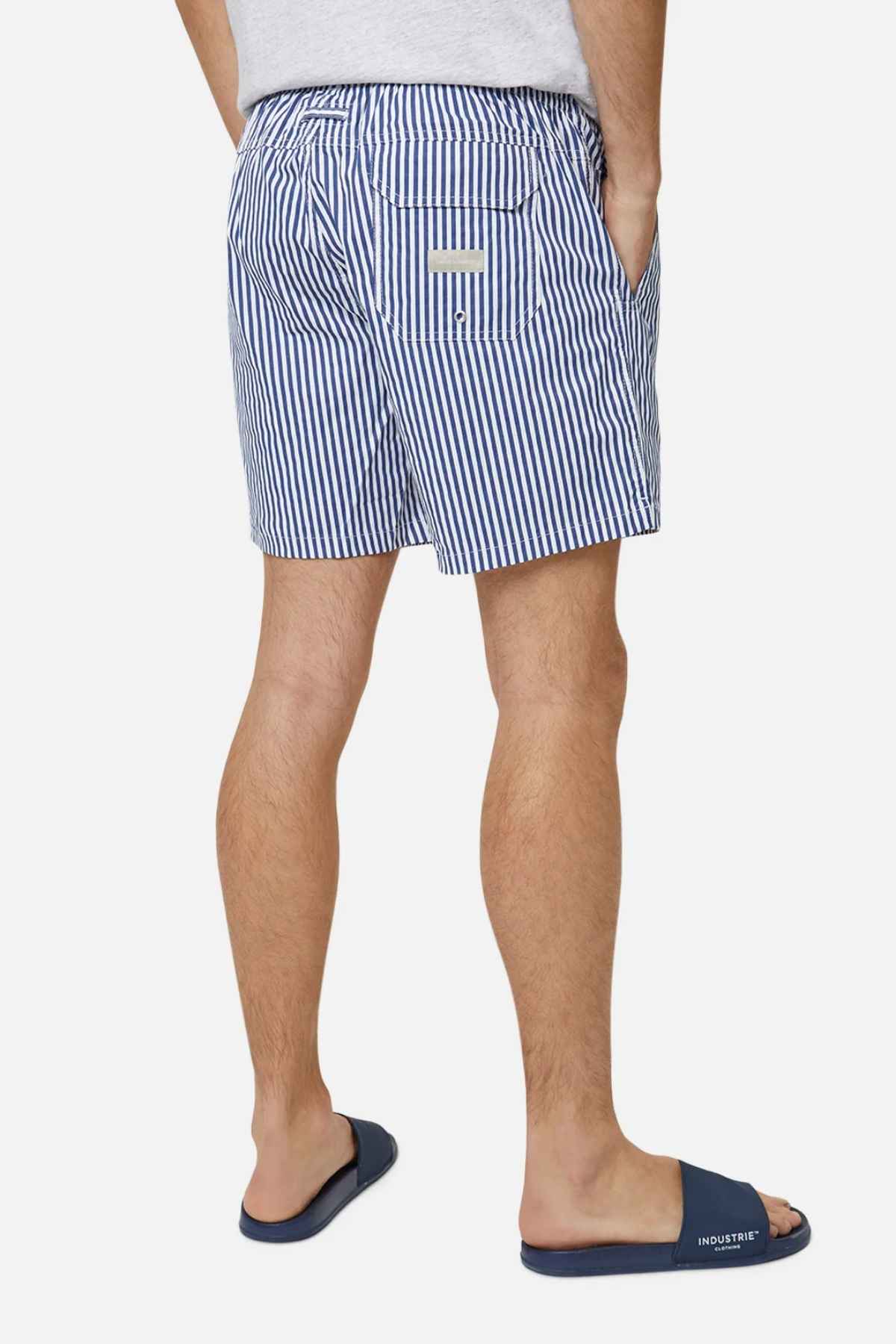 A man wearing Industrie The Princeton Bahama Short and navy blue slide sandals stands with his back to the camera. The comfortable fabric blend shorts include a small back pocket, set against a plain white background.