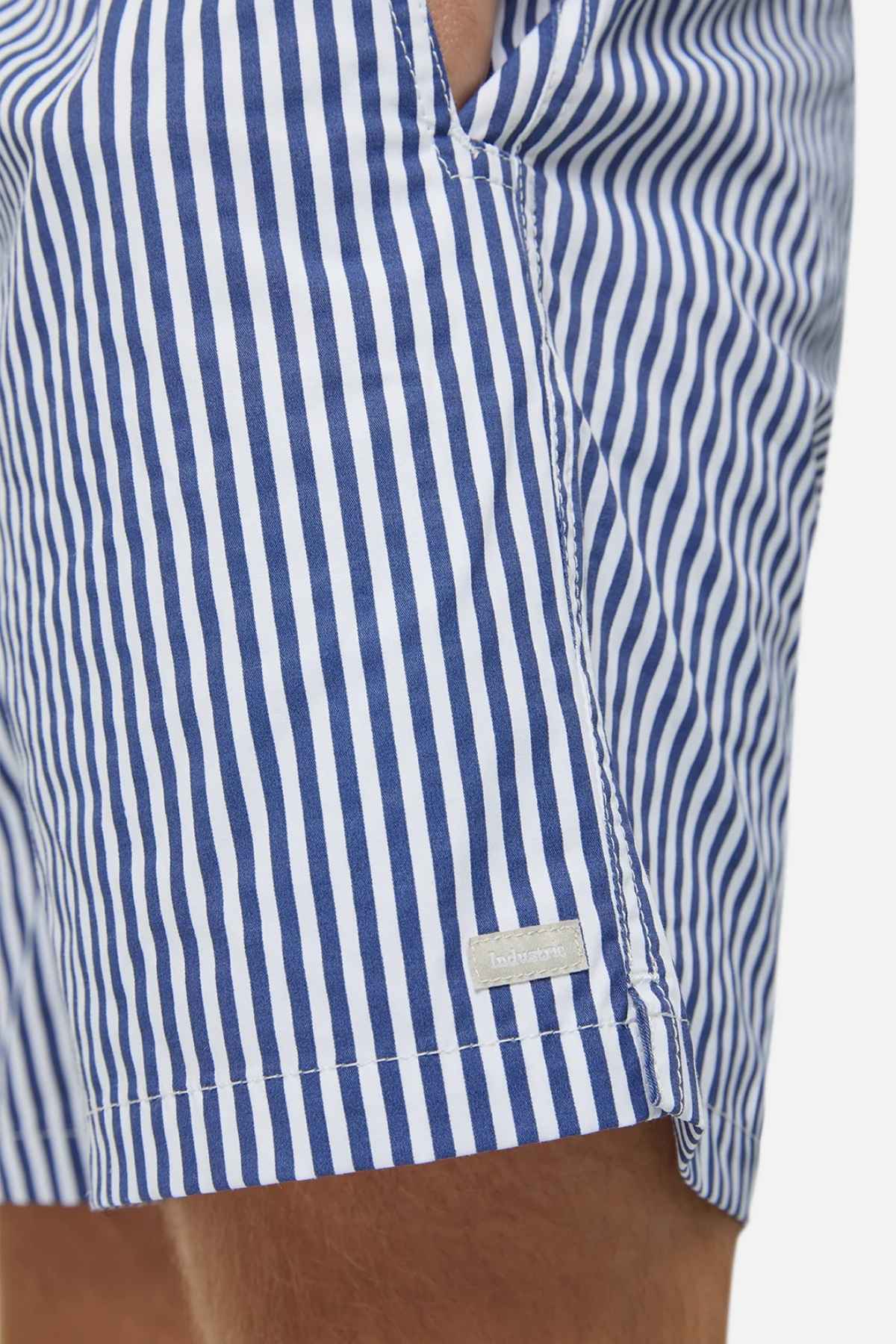 Close-up of a person wearing Industrie The Princeton Bahama Short, featuring a blue and white vertical stripe pattern. The comfortable fabric blend has a textured finish, highlighted by a small white label on the side seam. The background is plain and blurred.