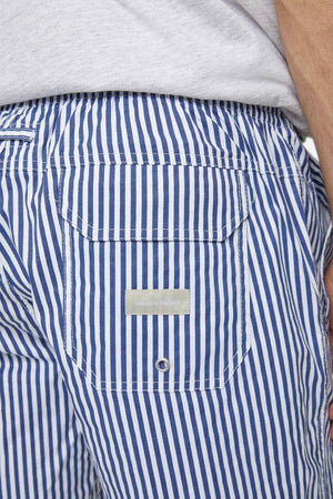 Close-up of someone wearing Industrie The Princeton Bahama Short, showcasing its blue and white stripes and handy back pocket. The tag indicating the comfortable fabric blend suggests premium quality. Their hand rests by their side, accompanied by a light-colored shirt visible at the top.