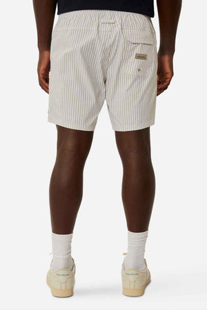A person dressed in Industrie The Princeton Bahama Shorts, which have cream-colored vertical stripes and a back pocket adorned with a small Industrie branding tag, stands against a plain white background. They pair the shorts with white socks and Reebok sneakers.