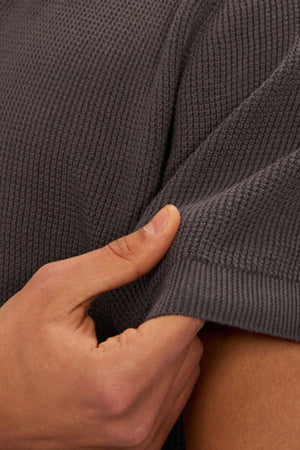 A close-up of a hand gently pulling the short sleeve of the Industrie The Ravenna Polo in dark gray, showcasing its textured knit fabric and stretch.