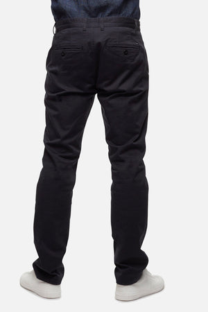 A person stands facing away, hands in pockets, wearing Industrie The Regular Cuba Chino Pant in a stretch cotton blend with a wider leg fit and white sneakers against a plain, light-colored background.
