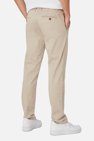A person stands with hands in pockets, sporting beige Industrie The Regular Cuba Chino Pant made from a stretch cotton blend and white sneakers. The chino pants, known for their wider leg fit, feature back pockets with button closures. The background is white.
