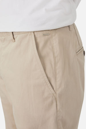Close-up view of a person wearing light beige Industrie The Regular Cuba Chino Pant and a white shirt. The pants feature a side pocket, a small label stitched on the seam near the pocket, and have a wider leg fit. The fabric appears to be made of stretch cotton blend or similar material.