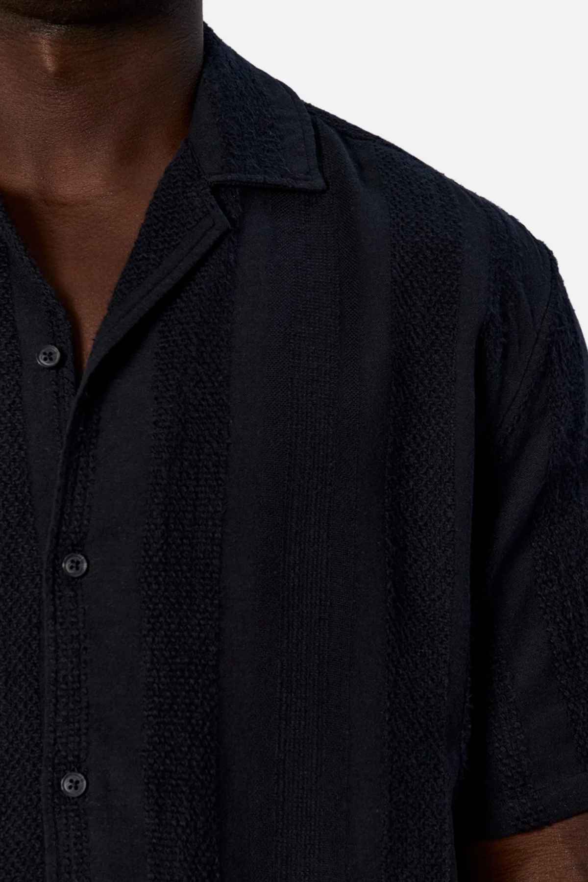 A person dressed in an Industrie The Salamanca Short Sleeve Shirt, crafted from a cotton-linen blend with a subtle textured pattern and notched collar, stands against a plain white background. Ideal for any summer wardrobe, the shirt is black and the individual's face is not visible.