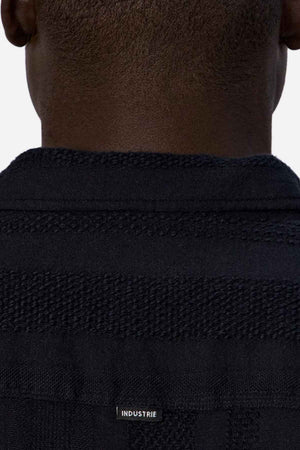 Close-up of the back of a person's neck and shoulders, wearing a textured black jacket with the word "INDUSTRIE" in small white letters on the back—a perfect addition to your summer wardrobe that pairs well with essentials like Industrie The Salamanca Short Sleeve Shirt.