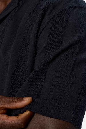 Close-up of a person wearing the Industrie The Salamanca Short Sleeve Shirt, crafted from a textured cotton-linen blend. Their hand gently pulls on the cuff of the short sleeve, highlighting its subtle vertical patterns. Ideal for your summer wardrobe, set against a plain white background.