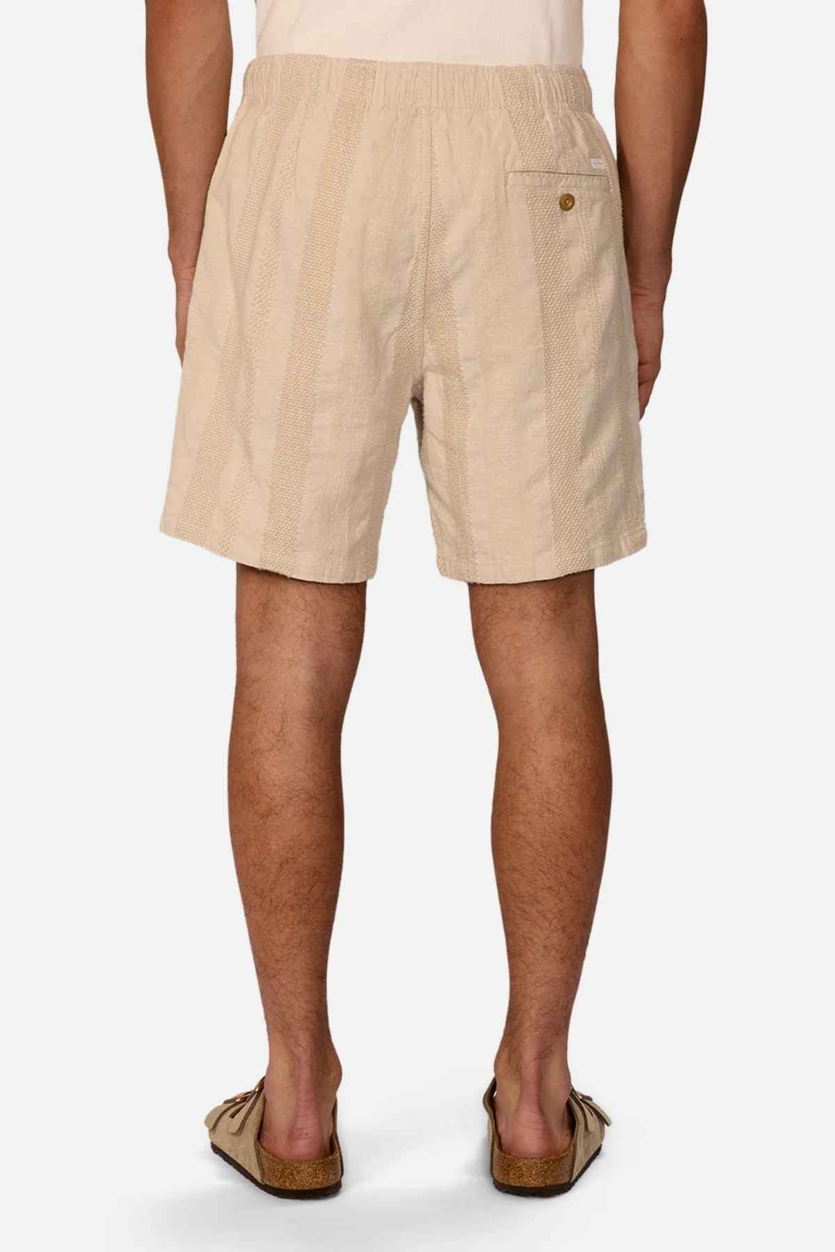 A person is shown from behind wearing a pair of Industrie The Salamanca Shorts in light beige with subtle vertical stripes, tailored fit, and a right side pocket, along with brown slip-on sandals. The shorts are crafted from a cotton linen blend, and the background is plain white.