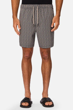 A man stands against a plain white background wearing Industrie The Savanna Swim Short, a black top, and black slide sandals.