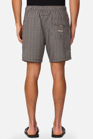 A person stands with their back to the camera, wearing Industrie The Savanna Swim Short with geometric print and back pocket, a black shirt, and black flip-flops. The quick-dry shorts stand out against a plain white background.