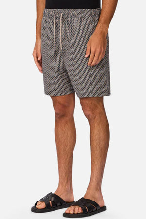 A man wearing Industrie The Savanna Swim Short with a black top and sandals stands against a plain light background, showing only his lower torso and legs.