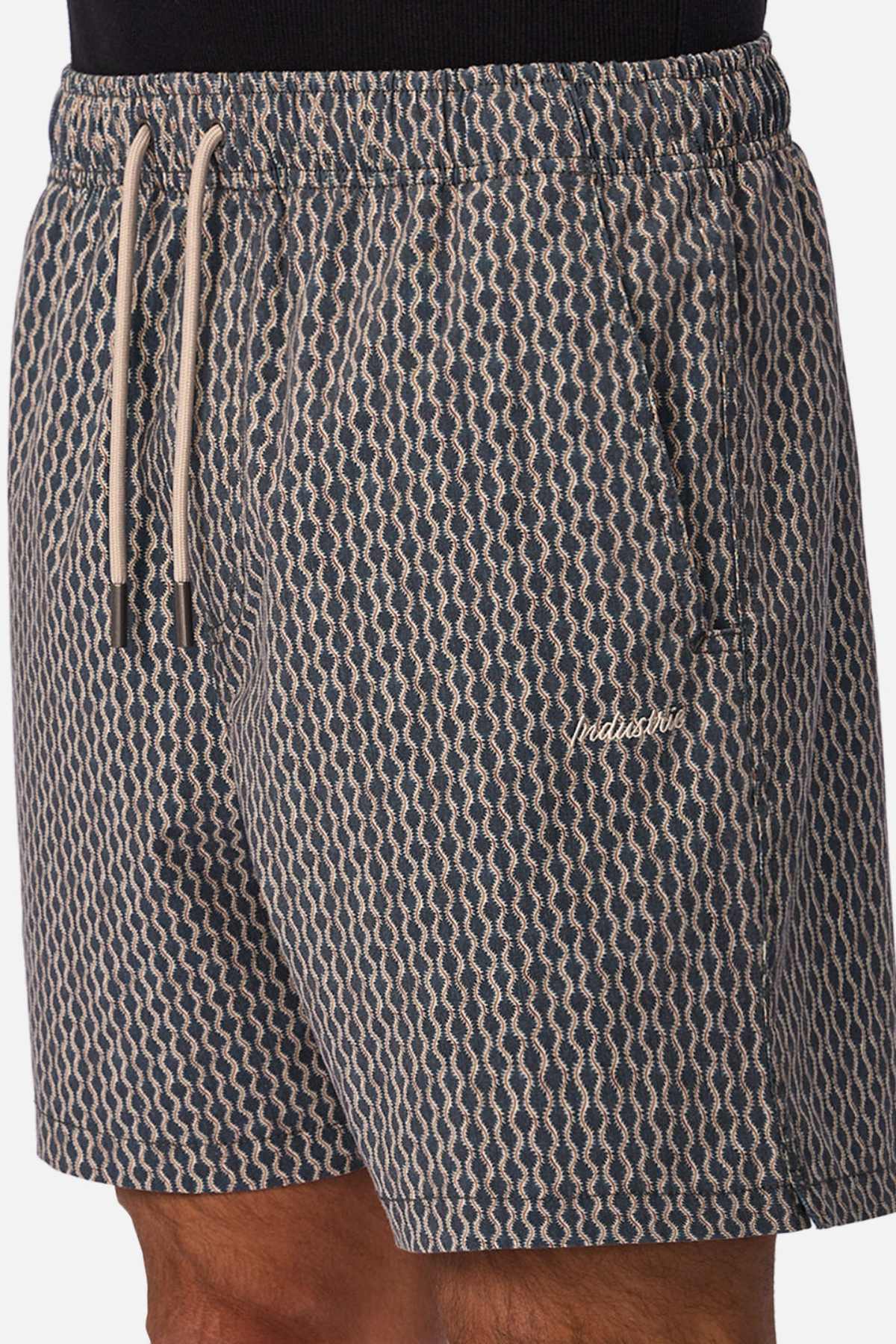 A person wearing Industrie The Savanna Swim Short in navy blue and beige pattern, featuring a white drawstring, quick-dry fabric, and a small embroidered logo on the left thigh.