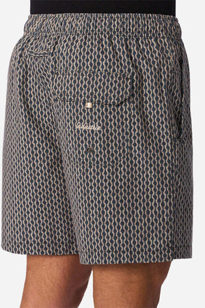 Wearing Industrie The Savanna Swim Short in blue and beige geometric print, quick-dry fabric, back pocket with button flap and embroidered logo. Photo shows side-rear waist to mid-thigh view.