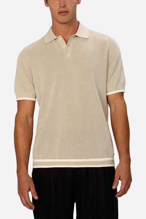 A person is dressed in a beige Industrie The Serrano Polo, crafted entirely from cotton and showcasing a cable knit design along with short sleeves. It's paired with black pants, and the shirt's textured pattern is highlighted against the plain white backdrop.