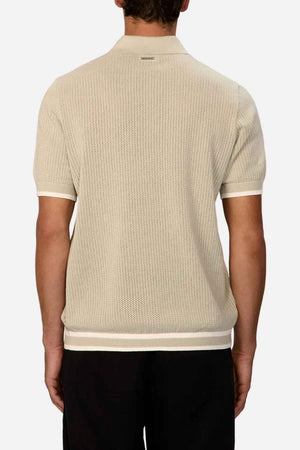 The image depicts a person from behind wearing the Industrie The Serrano Polo, a light beige short-sleeved polo shirt made from 100% cotton. It is perfectly complemented by black pants. The shirt showcases a textured pattern with ribbed cuffs and a classic collar, providing an effortlessly stylish appearance.