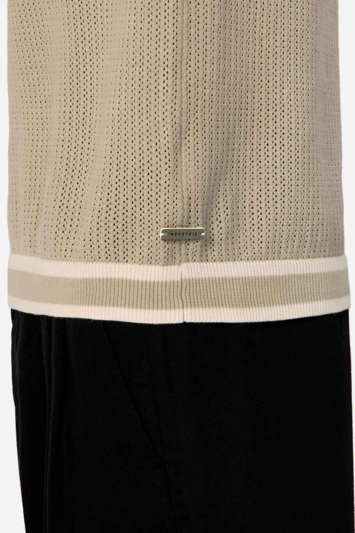 A close-up of the Industrie The Serrano Polo showcases its beige mesh jersey design, highlighted by a small rectangular silver tag on the hem. Made from 100% cotton, this top is layered over black pants and features a contrasting white stripe at the bottom hem.