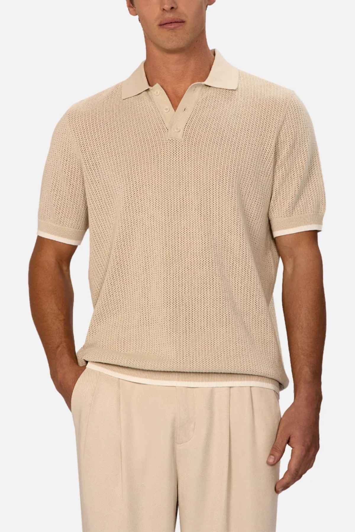A man is wearing The Serrano Polo by Industrie, a chic beige cable knit polo shirt, paired with matching pants. The shirt features a stylish collar and short sleeves, as his left hand rests casually in his pocket against the plain white background.