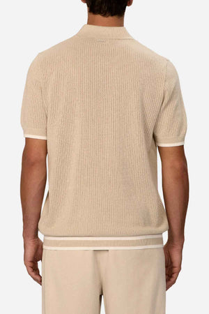 Rear view of a person wearing a beige short-sleeved knit shirt and matching pants, standing against a neutral background. The shirt, similar to Industrie The Serrano Polo, showcases a textured pattern with white trim on the cuffs and hem, adding subtle sophistication.