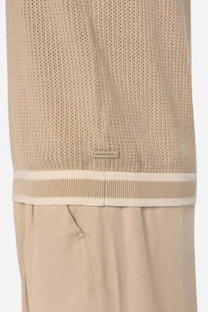 Close-up of a beige cable knit garment with a perforated design, featuring white and beige trim along the lower edge adorned with the word "INDUSTRIE" in small letters. Below, matching beige pants enhance the chic look of the Industrie The Serrano Polo ensemble.