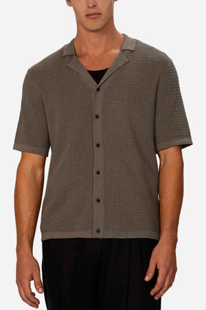 A man wears the Industrie The Sevona Short Sleeve Shirt, featuring a textured, short-sleeve, button-up design in a neutral grey hue over a black undershirt. Offering a relaxed fit perfect for any summer wardrobe, he pairs it with dark pants against a plain white background.