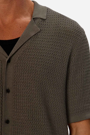 Close-up of a person wearing the Industrie The Sevona Short Sleeve Shirt, a textured, knitted olive green short-sleeve button-up with a relaxed fit over a black undershirt. Perfect for any summer wardrobe, it features a woven pattern and spread collar with black buttons, focusing on the upper torso area.