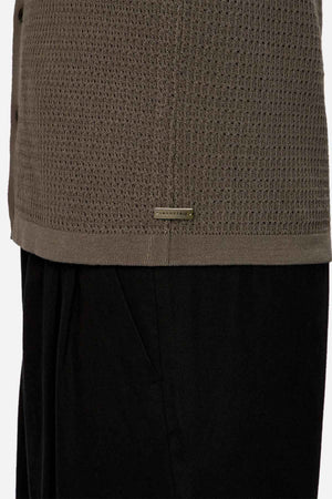 A close-up showcases someone wearing the textured olive green Industrie The Sevona Short Sleeve Shirt, an ideal addition to a summer wardrobe with its relaxed fit and subtle rectangular tag near the hem. It's stylishly paired with black pants against a plain background.