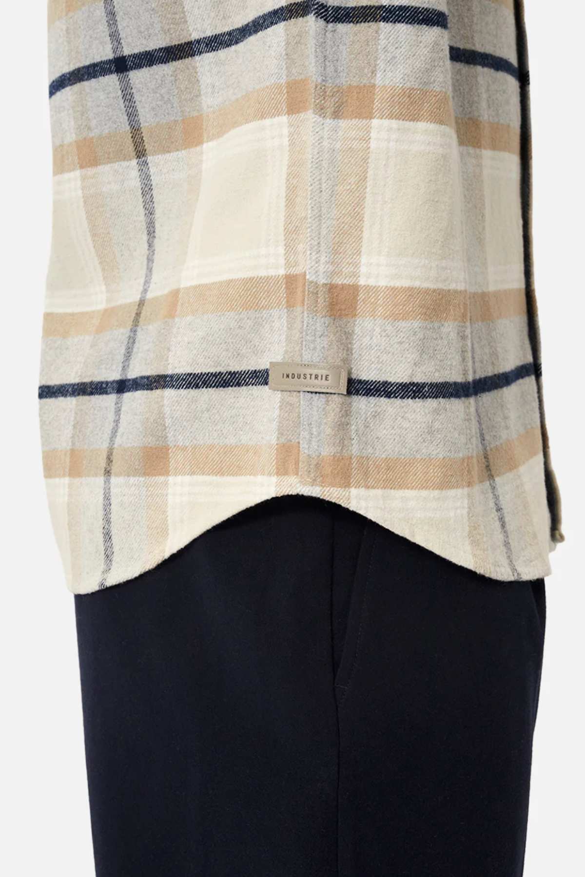 A close-up shows someone in black pants wearing Industrie The Somerville Shirt with a cream, tan, and navy plaid design. The shirt features the Industrie branding label against a plain white background.