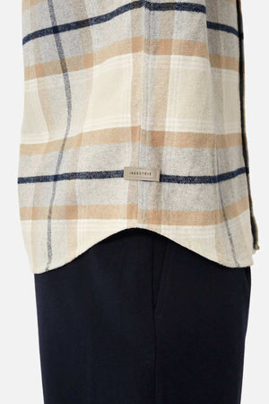 A close-up shows someone in black pants wearing Industrie The Somerville Shirt with a cream, tan, and navy plaid design. The shirt features the Industrie branding label against a plain white background.