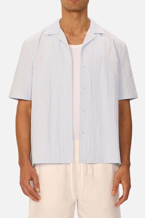 A man wears the Industrie The Sorrento Short Sleeve Shirt in light blue and white pinstripes over a white tee with light pants. The image is cropped from just below his shoulders to above his knees.