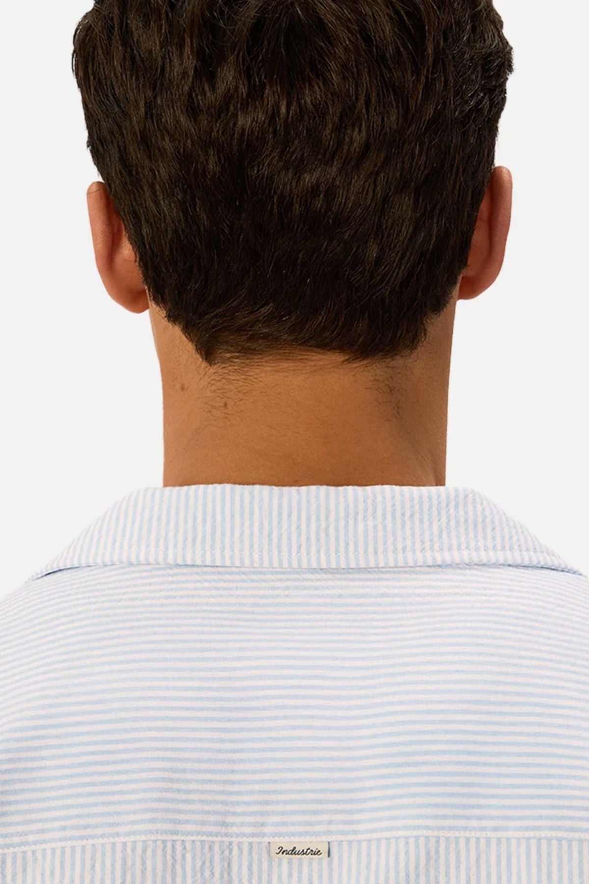 The image shows the back view of a person with short dark hair wearing the Industrie The Sorrento Short Sleeve Shirt, which features light blue and white pinstripes. The background is plain white.