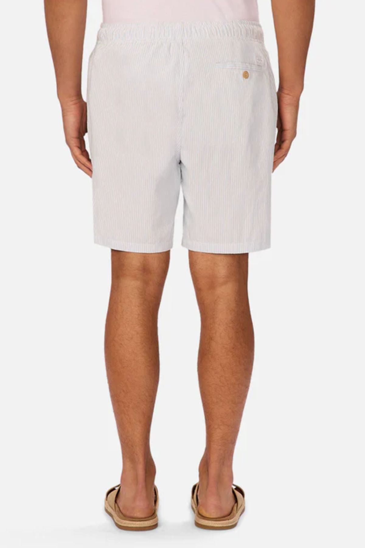A person stands facing away, wearing Industrie The Sorrento Short in light blue and white pinstripes with an elastic waistband, a single buttoned back pocket, tan sandals, and a white shirt.