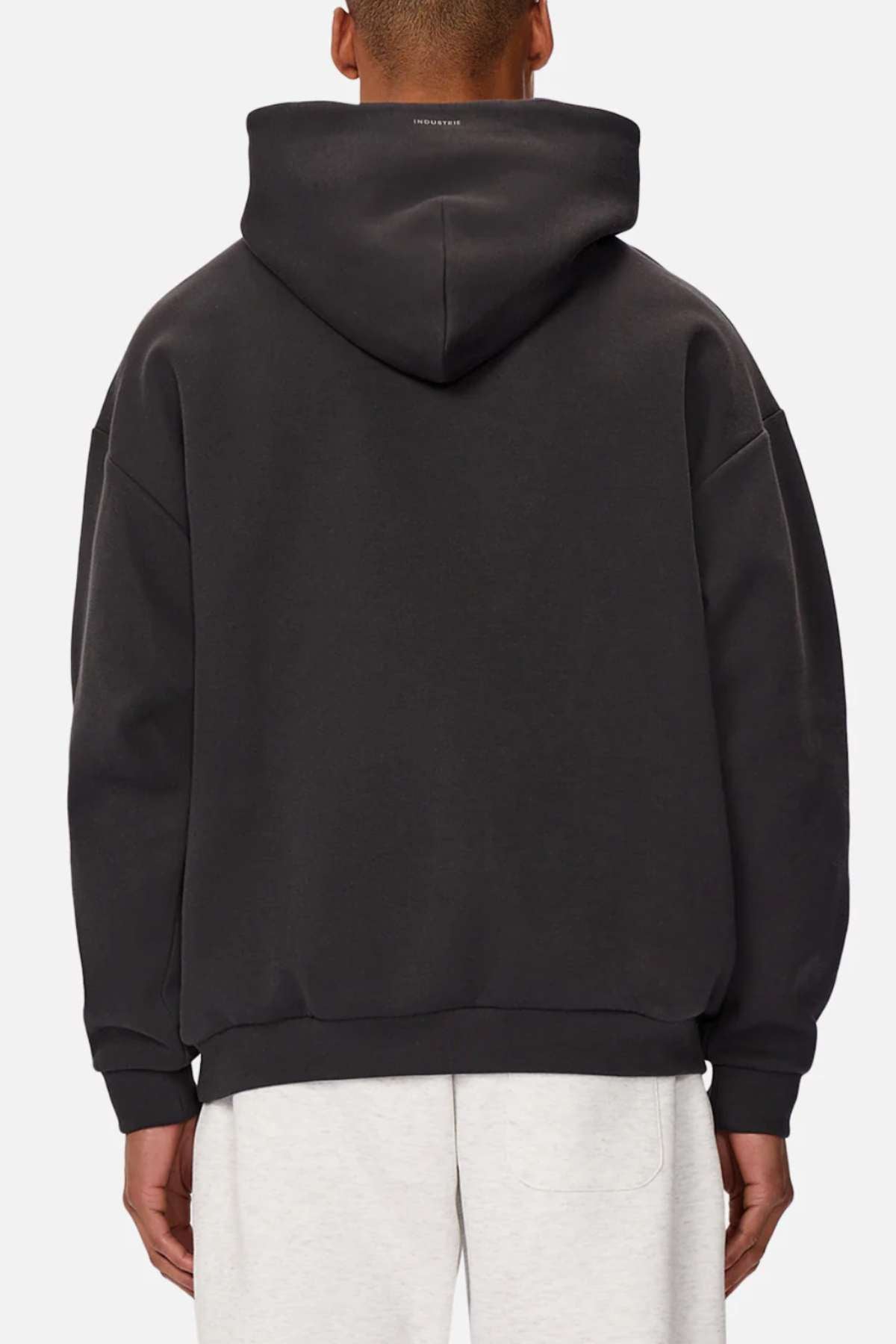 A person is seen from the back wearing Industrie The Tech Del Sur Hood in black, crafted with advanced athletic fabrications, paired with light gray pants. It features a slightly oversized fit and a small logo at the top on a plain white background.