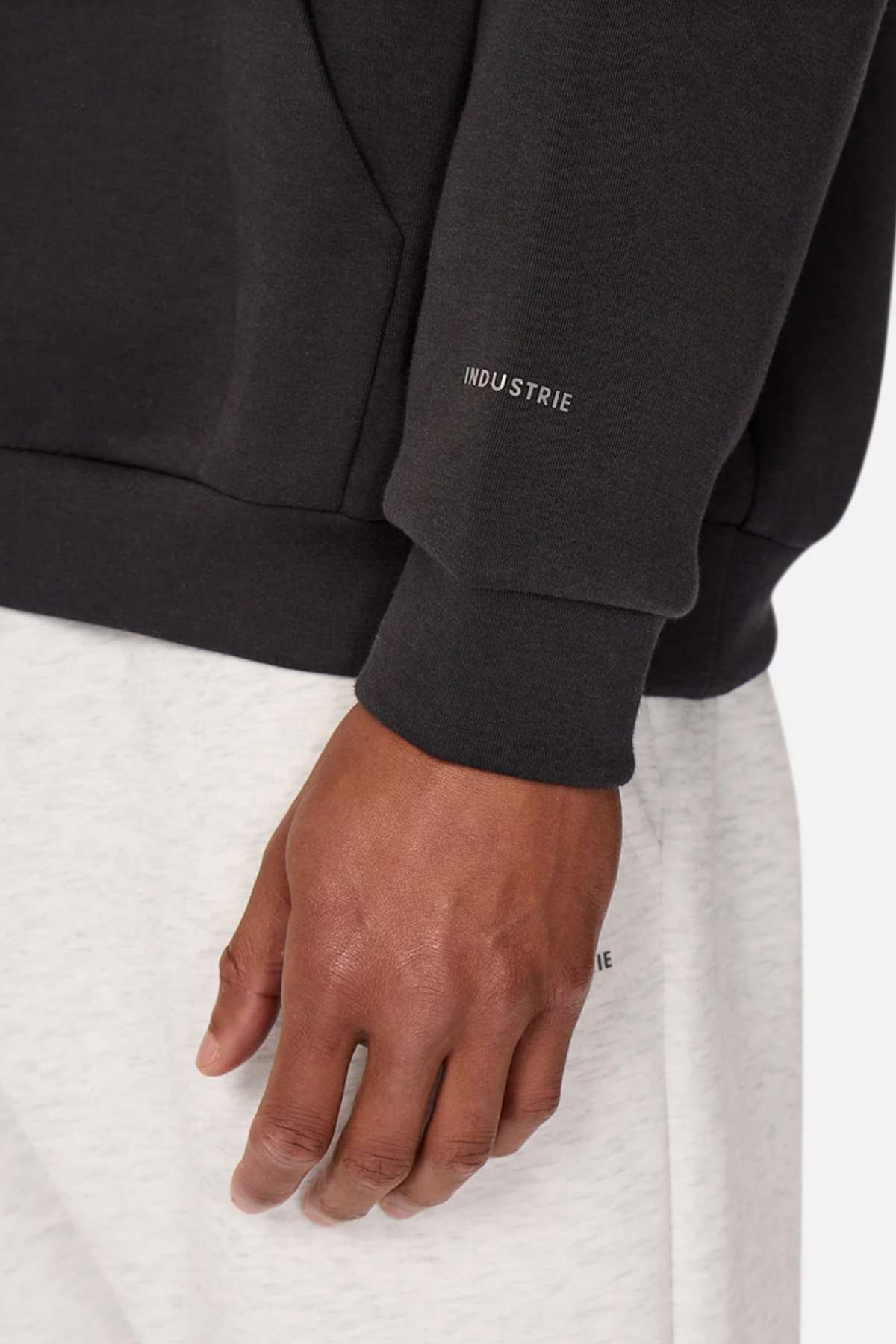 Close-up of a person wearing the Industrie The Tech Del Sur Hood, crafted from advanced athletic fabric, with "INDUSTRIE" on the sleeve, paired with light-colored pants. They stand against a neutral background, exuding modernity as their hand rests casually by their side.