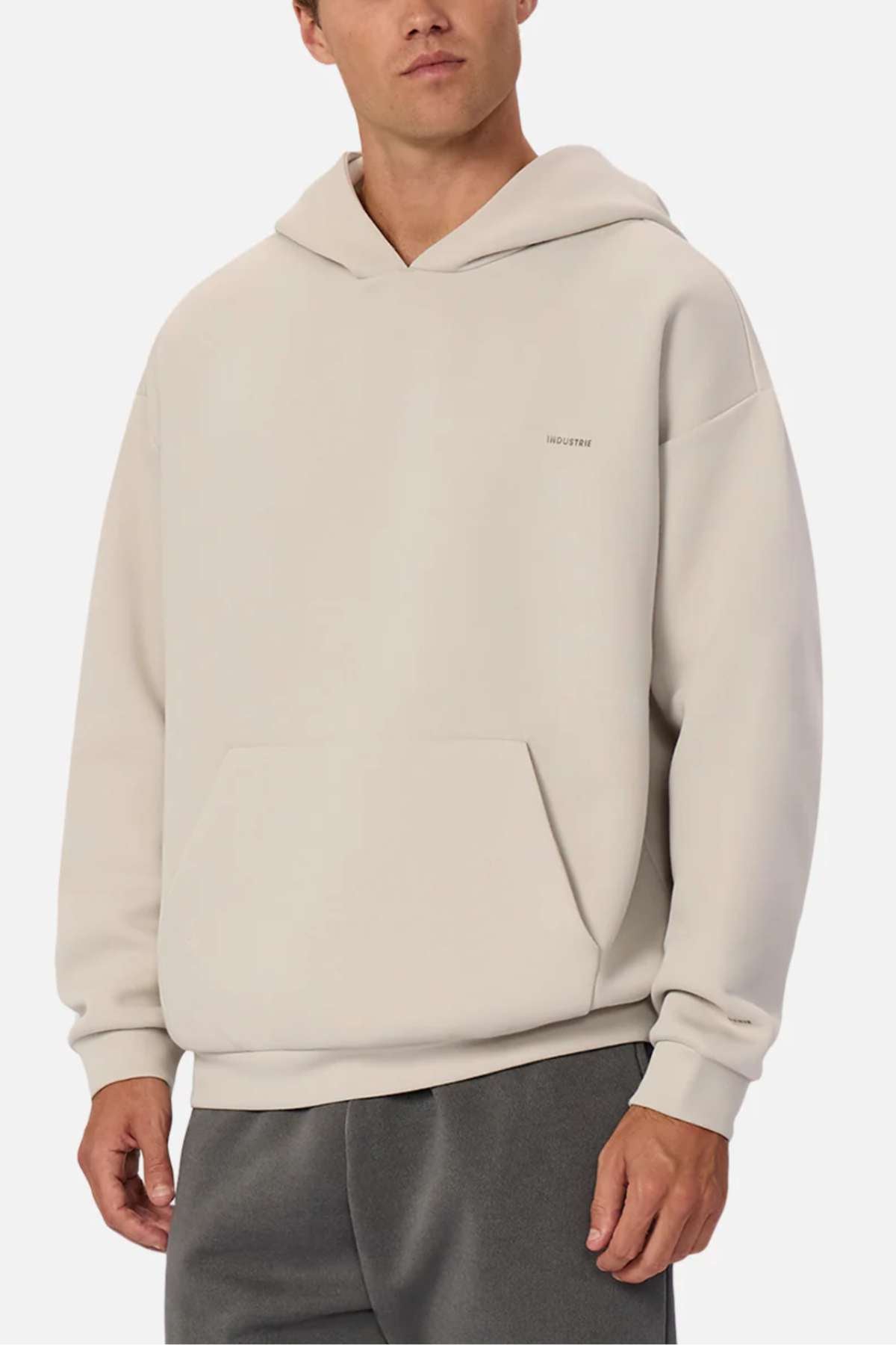 A man wears the Industrie The Tech Del Sur Hood in light beige, featuring a front pocket and subtle chest logo, paired with dark gray athletic pants, standing against a plain white background.