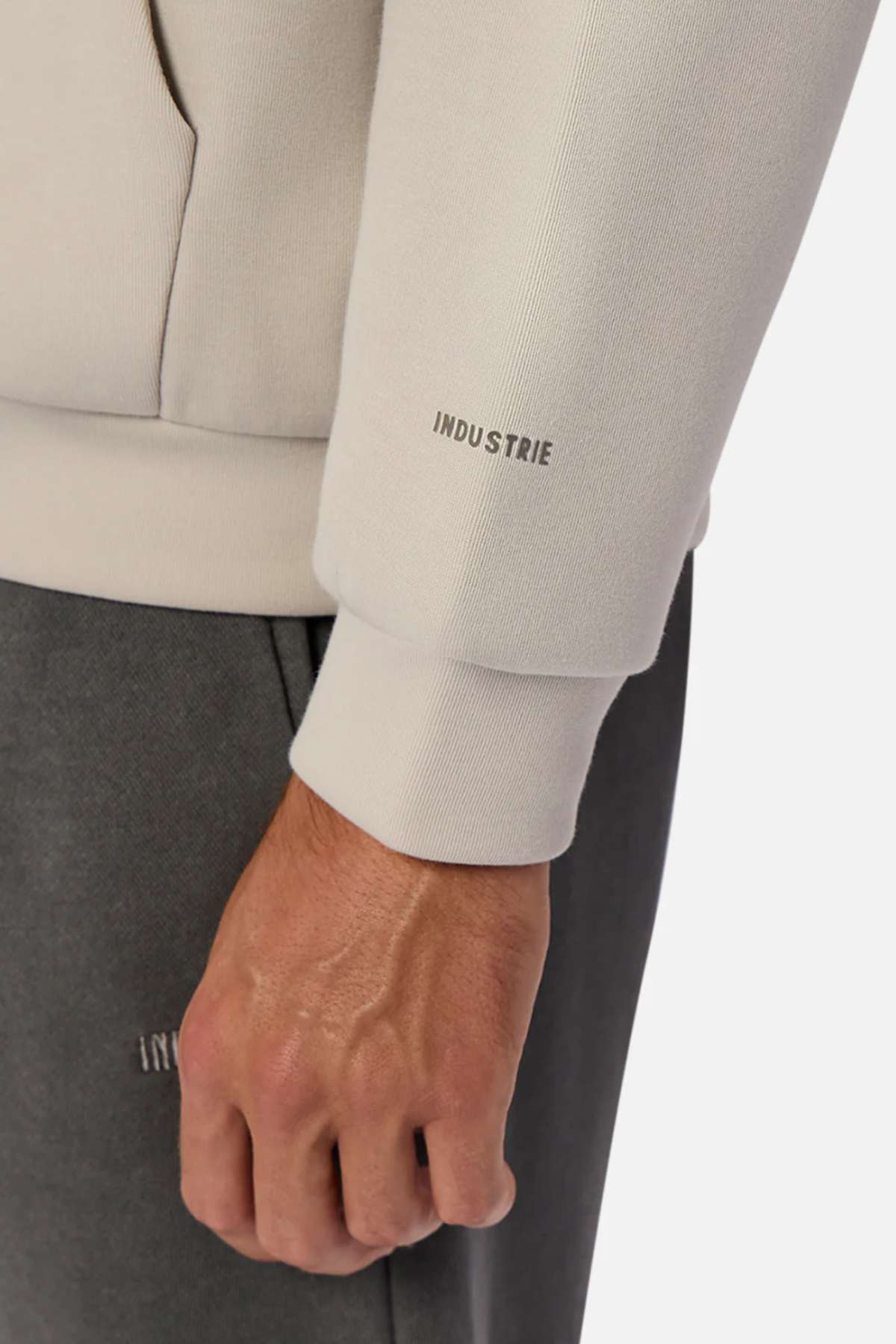 A close-up of a hand and wrist wearing the Industrie The Tech Del Sur Hood in light beige, showing the INDUSTRIE logo on the cuff, paired with dark gray pants featuring matching branding.