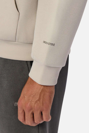 A close-up of a hand and wrist wearing the Industrie The Tech Del Sur Hood in light beige, showing the INDUSTRIE logo on the cuff, paired with dark gray pants featuring matching branding.