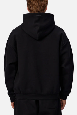 A person with short, wavy brown hair is seen from behind wearing black pants and an Industrie The Tech Del Sur Hoodie in black, hood up, with small white text visible on the back below the hood.