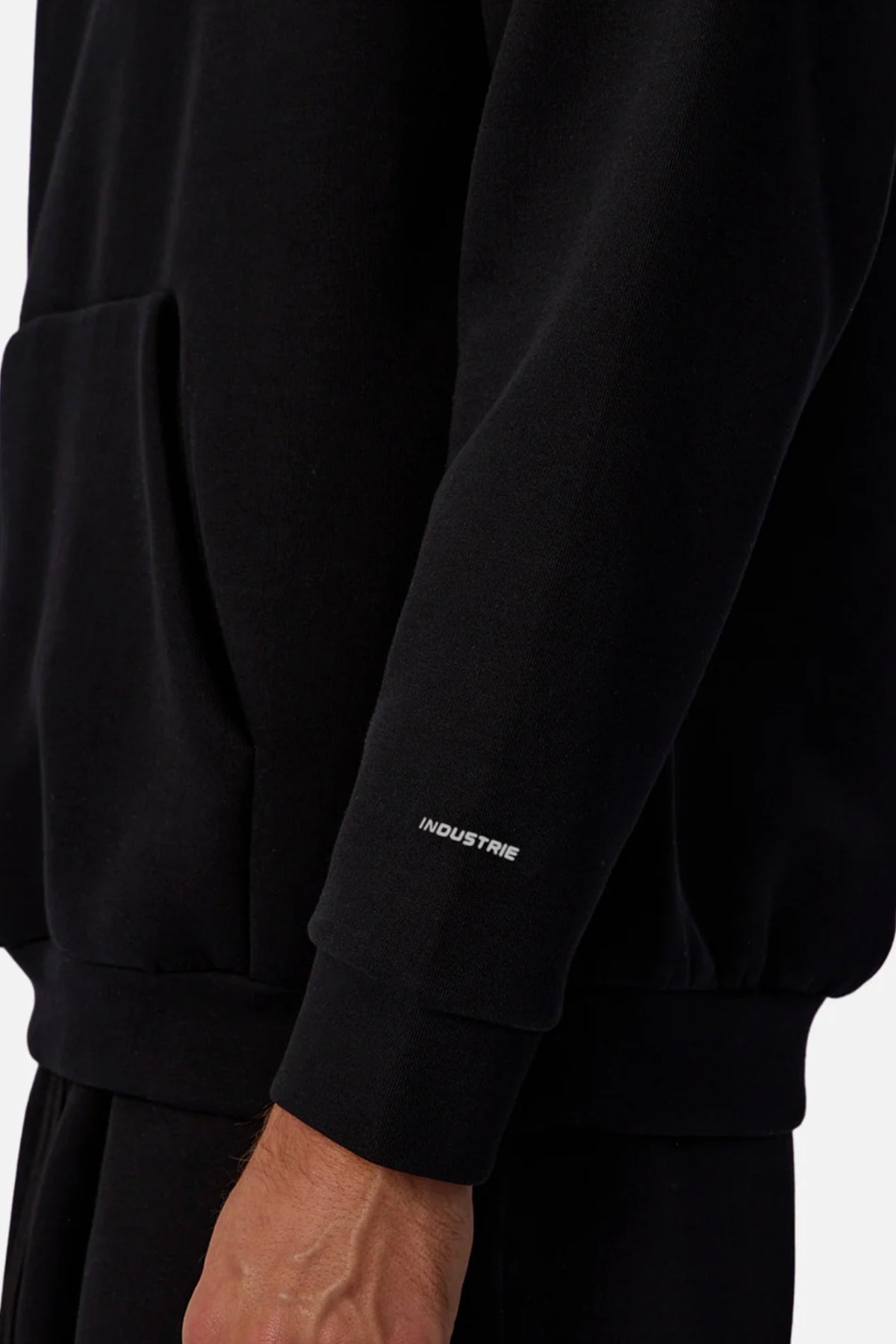 A close-up shows a person in the Industrie The Tech Del Sur Hoodie, which has "INDUSTRIE" in small white letters near the sleeve wrist, a front pocket, and the wearerβs hand partly visible.