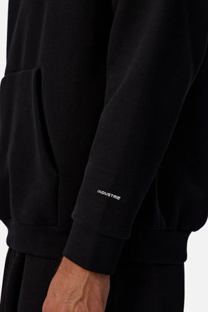 A close-up shows a person in the Industrie The Tech Del Sur Hoodie, which has "INDUSTRIE" in small white letters near the sleeve wrist, a front pocket, and the wearerβs hand partly visible.