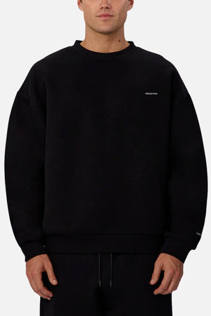 A person wears the loose-fit black Industrie The Tech Del Sur Sweat, featuring small white text on the left chest and wrist, with black pants, standing against a plain light background.