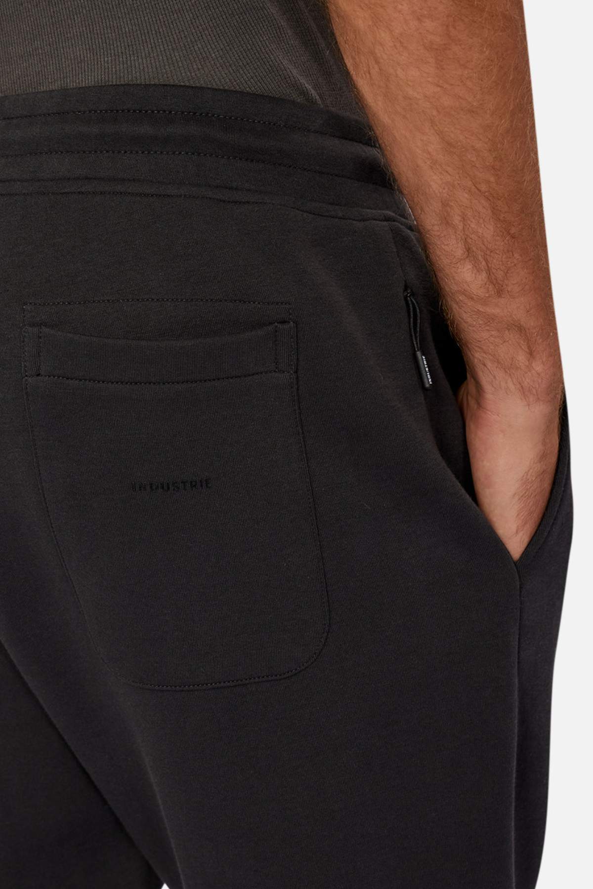 Close-up of someone wearing Industrie The Tech Del Sur Track Pant with dark athletic material, a visible back pocket, and their right hand in the front pocket. The fabric, featuring an elastic waistband, looks soft against a neutral background.
