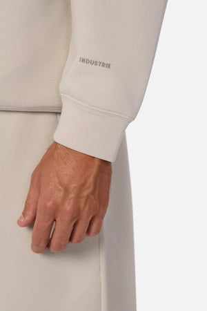 A close-up of a hand wearing a light beige Industrie The Tech Staunton Track Top shows "INDUSTRIE" printed near the wrist. Set against a white background, the design is sleekly highlighted with practical zip-up side pockets.