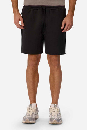 A person is standing, wearing Industrie The Tech Track Short with an elasticated drawstring waist and a gray T-shirt. They have white sneakers with gray details. The photo captures them from the knees to the shoulders against a plain background, offering an athletic feel.
