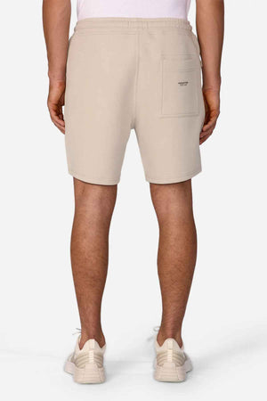 A person wearing The Tech Track Short by Industrie, featuring a versatile design in beige with the INCLINE logo on the back pocket, stands with white sneakers, facing away from the camera.