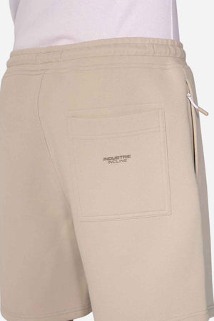 A person wearing Industrie The Tech Track Short showcases its versatile design in beige, complete with a visible pocket and small zipper detail. The soft, athletic material displays the "INDUSTRIE INCLINE" logo on the pocket against a plain white background.