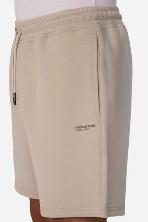 A person is wearing The Tech Track Short in beige, featuring a drawstring waist for adjustable comfort. These shorts, crafted from athletic material, display a compact "Industrie INCLINE" logo on the left side. The dynamic and versatile design is showcased with a white shirt, highlighting the side view of these sporty shorts.