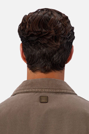 A person with short, wavy brown hair is seen from behind, wearing the Industrie The Tennesse Jacket—a men's cotton jacket featuring a stitched patch on the collar. The background is plain and light-colored.