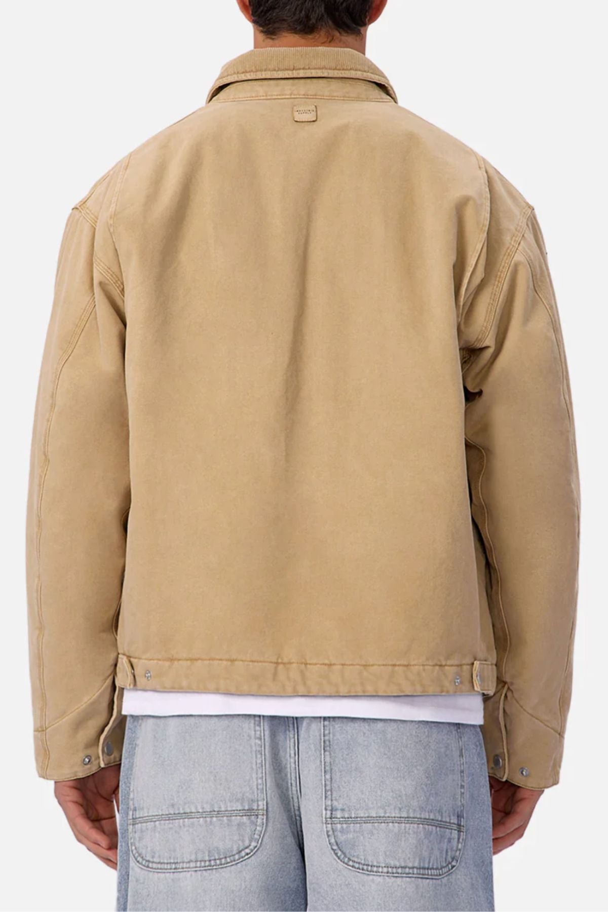 A person with short dark hair is seen from the back wearing the Industrie The Tennesse Jacket, a 100% cotton men’s casual beige waist-length jacket with visible stitching and metal waistband buttons, paired with light blue denim jeans.